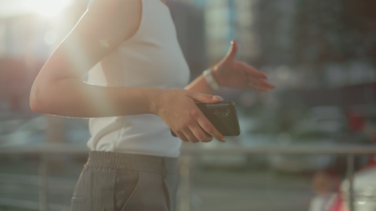 Close side view of lady in armless top holding phone dancing under golden sunlight, hand motion expressive with urban blur background adding warm ambiance and soft glow around subject