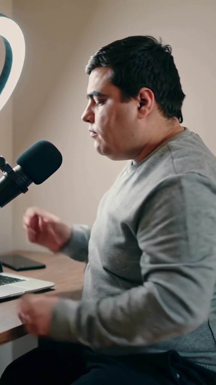 Vertikální video: Muž v šedé košili se opírá, mluví do mikrofonu a prohlíží si notebook doma pod kruhovým světlem.