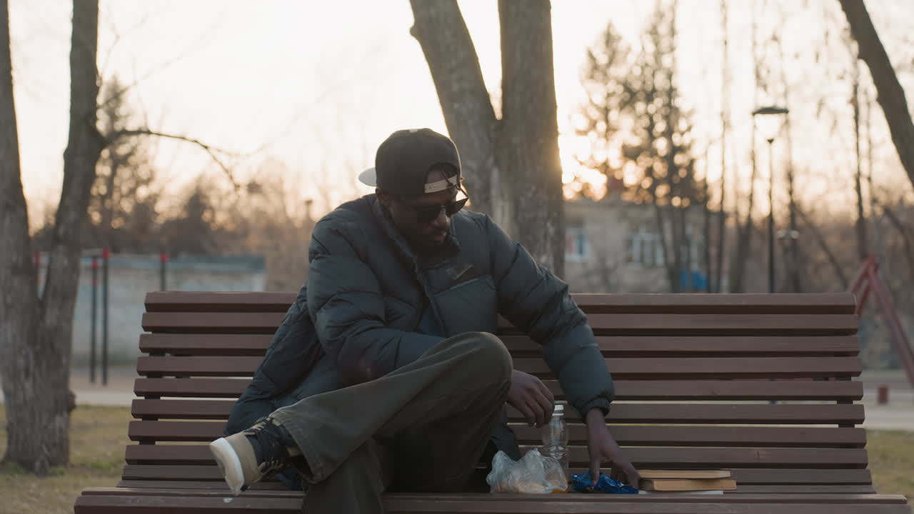 Man in sunglasses and cap sits on bench at sunset holding open bottle, puffer jacket zipped, calm expression, blurred park background with trees and buildings