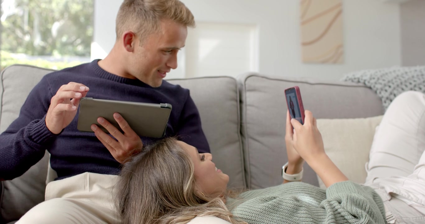 Couple on gray sofa, woman sharing smartphone screen while man leaning, tapping tablet, smiling