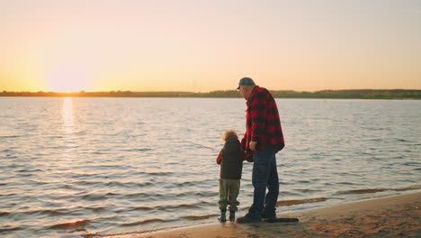 El-Abuelo-Está-Enseñando-A-Su-Nieto-Pequeño-A-Pescar-Con-Una-Caña-De-Pescar,-Un-Anciano-Y-Un-Niño-Pequeño-En-La-Orilla-Arenosa-Del-Río.