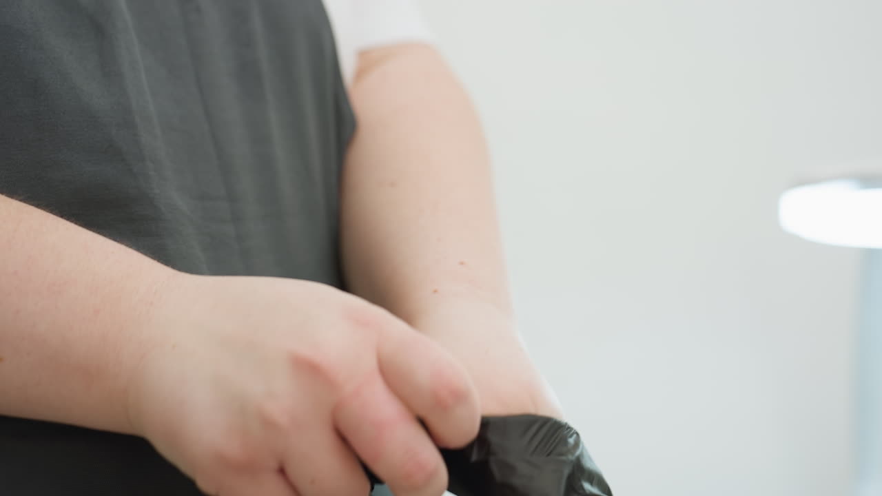 Close-up view of beautician wearing black glove over hand while dressed in dark apron, preparing for salon session in clean white space with blurred background and lamp for lighting focus