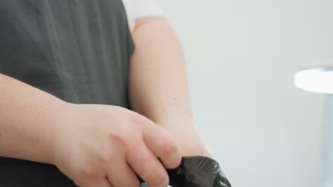 close-up view of beautician wearing black glove over hand while dressed in dark apron, preparing for salon session in clean white space with blurred background and lamp for lighting focus