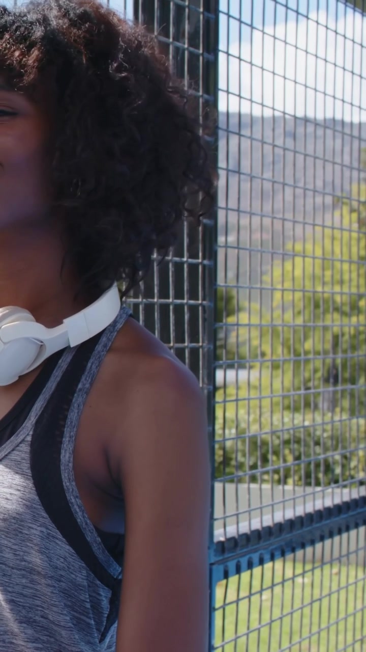 Vertical video: Turning woman in grey tank lifting gaze by fence after tap, headphones, strap