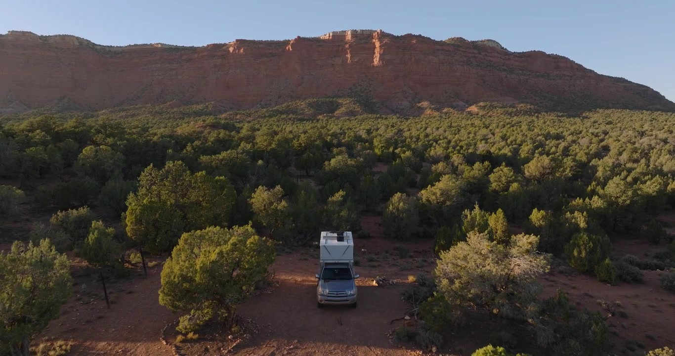 Furgoneta camper estacionada entre enebros verdes del desierto bajo una meseta de arenisca roja cerca de Kanab, Utah, toma de dron en movimiento al atardecer