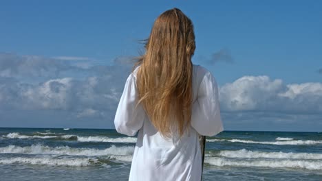 a woman standing on the beach, the wind blowing through her hair and her dress, with the waves crashing in the background