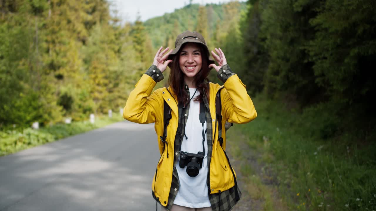 Portrait of a tourist girl in special hiking clothes, along with a camera in a yellow jacket, who looks into the camera against the backdrop of a mountain forest