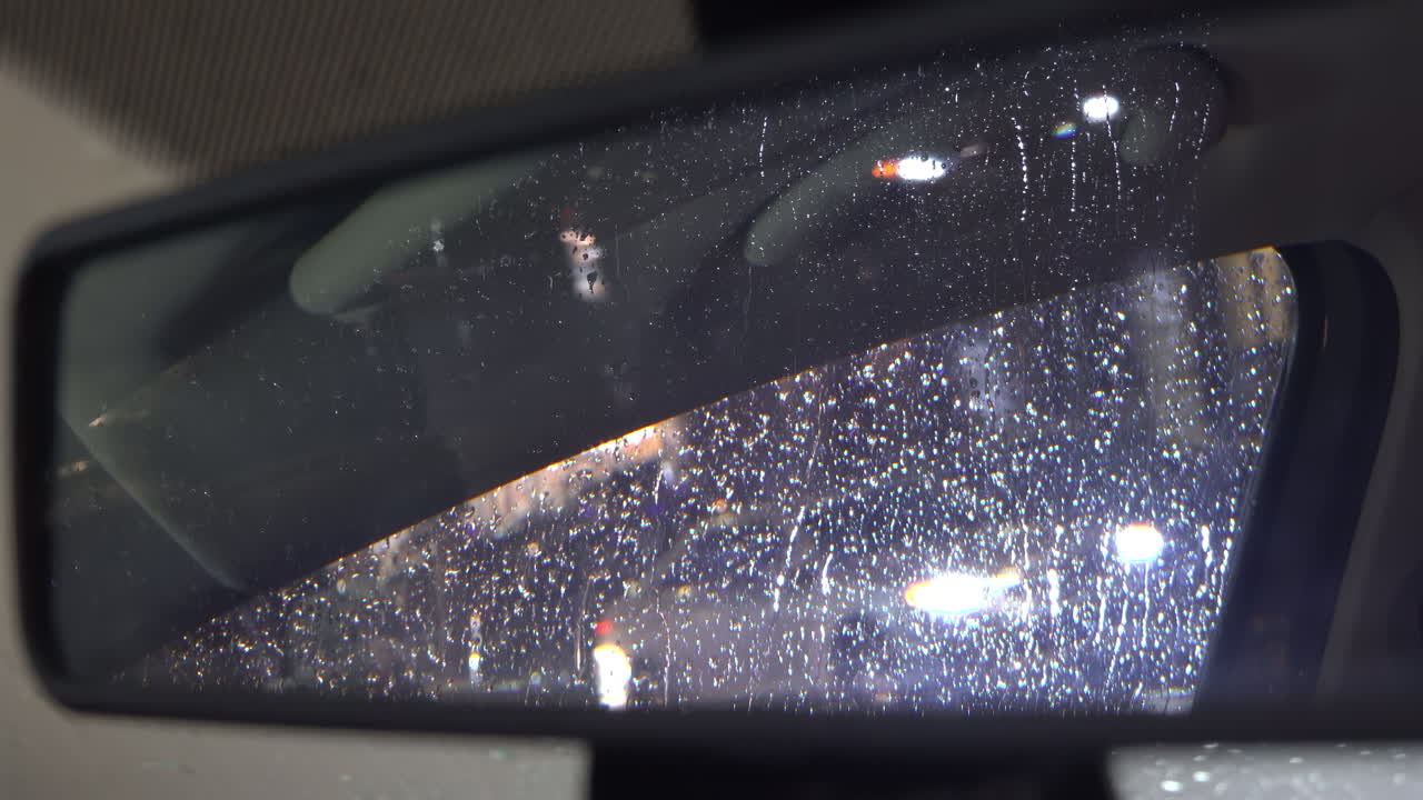 Rain dripping down the window of a moving car in the evening reflected in the rearview mirror
