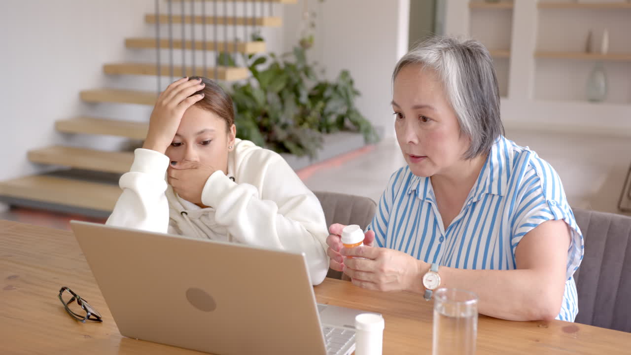 Sitting at table, asian grandmother showing pill bottle to girl using laptop
