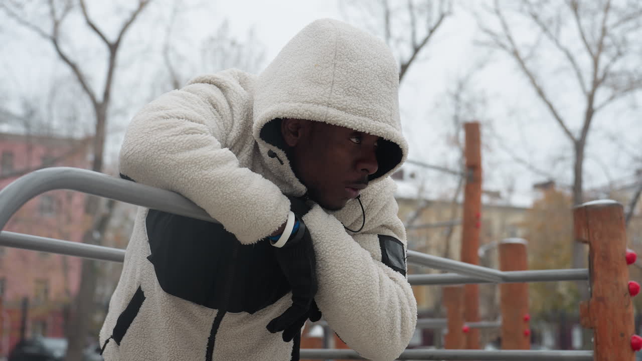 Young man in white sweater resting on iron bar, breathing heavily during intense workout in winter, he wears gloves and a hooded jacket, exhaling visible cold breath in outdoor urban park setting