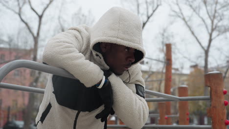 young man in white sweater resting on iron bar, breathing heavily during intense workout in winter, he wears gloves and a hooded jacket, exhaling visible cold breath in outdoor urban park setting