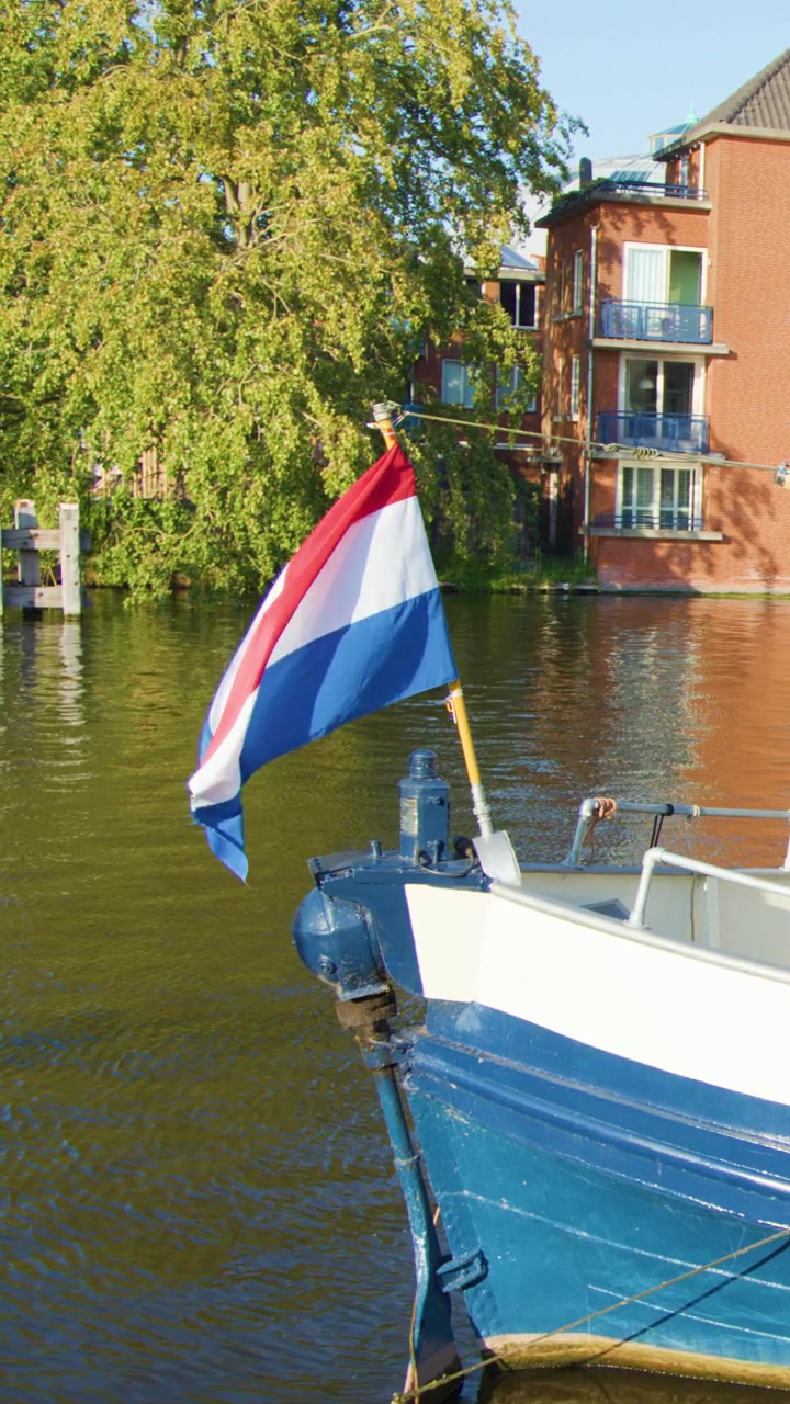 Traditionelles niederländisches Boot mit Nationalflagge gleitet entlang des sonnenbeschienenen Haarlem-Kanals vorbei an modernen Gebäuden.