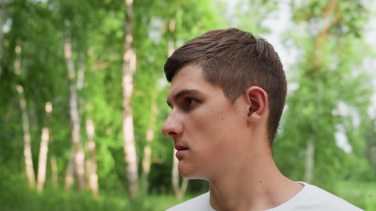 Medium view of young man wearing white shirt wiping sweat from face with hand while standing under summer sunlight in green forest background, showing natural movement, and casual emotion outdoors