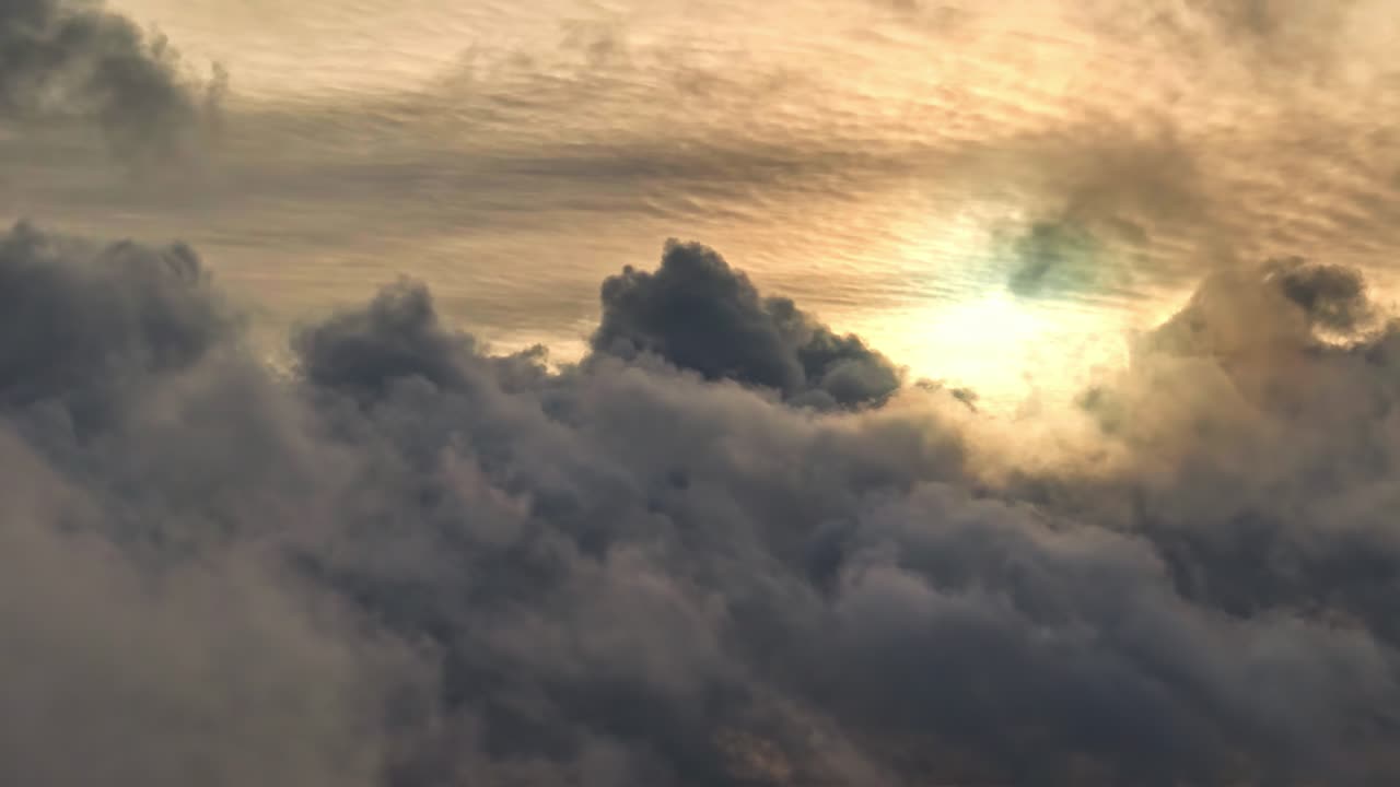 Vista aerea del sole che si fa strada attraverso uno strato denso di nuvole temporalesche scure, proiettando una luce dorata sulle cime delle nuvole. Contrasto drammatico tra ombra e bagliore caldo all'ora d'oro.