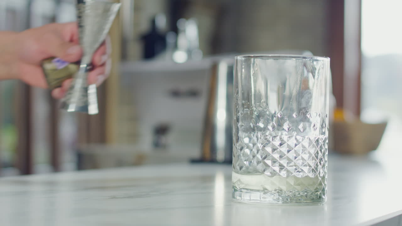 Close-up of a bartender's hand placing a glass while holding a jigger in the out-of-focus background