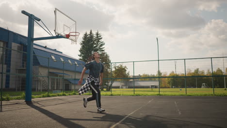 man in outdoor court throws up volleyball preparing for a slam with background showing a basketball hoop, fence, and a green field, athletic action shot in a sport environment