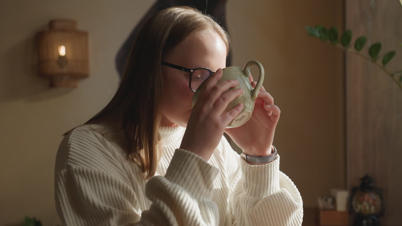 Content woman in white sweater holds warm cup near face and inhales aroma of coffee while seated near window, sunlight casting soft shadows on wall with potted plant and cozy interior details