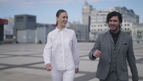 confident man and woman talking walking on city square in slow motion leaving. portrait of confident elegant intelligent successful caucasian coworkers strolling downtown outdoors chatting.