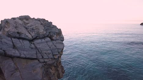 Drohnenschuss-Aus-Der-Luft,-Der-Während-Der-Blauen-Stunde-In-Griechenland-Neben-Einem-Großen-Felsen-Im-Ruhigen-Meer-Fliegt