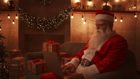 Papá-Noel-Está-Sentado-En-Un-Cómodo-Sofá-Suave-Sosteniendo-Una-Computadora-Portátil-En-Su-Regazo-Y-Escribiendo-En-Ella.-Hermoso-Salón-Decorado-Para-Una-Reunión-Feliz-Navidad-Y-Próspero-Año-Nuevo.-Imágenes-4k-De-Alta-Calidad