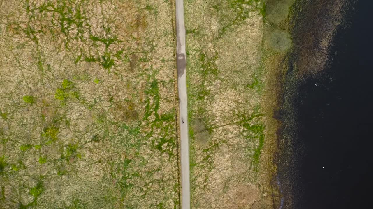 Top down aerial drone footage view of a person walking on a desolate rural paved road in Scotland british green moss covered nature besides a large lake that is dark colored during beautiful day time.