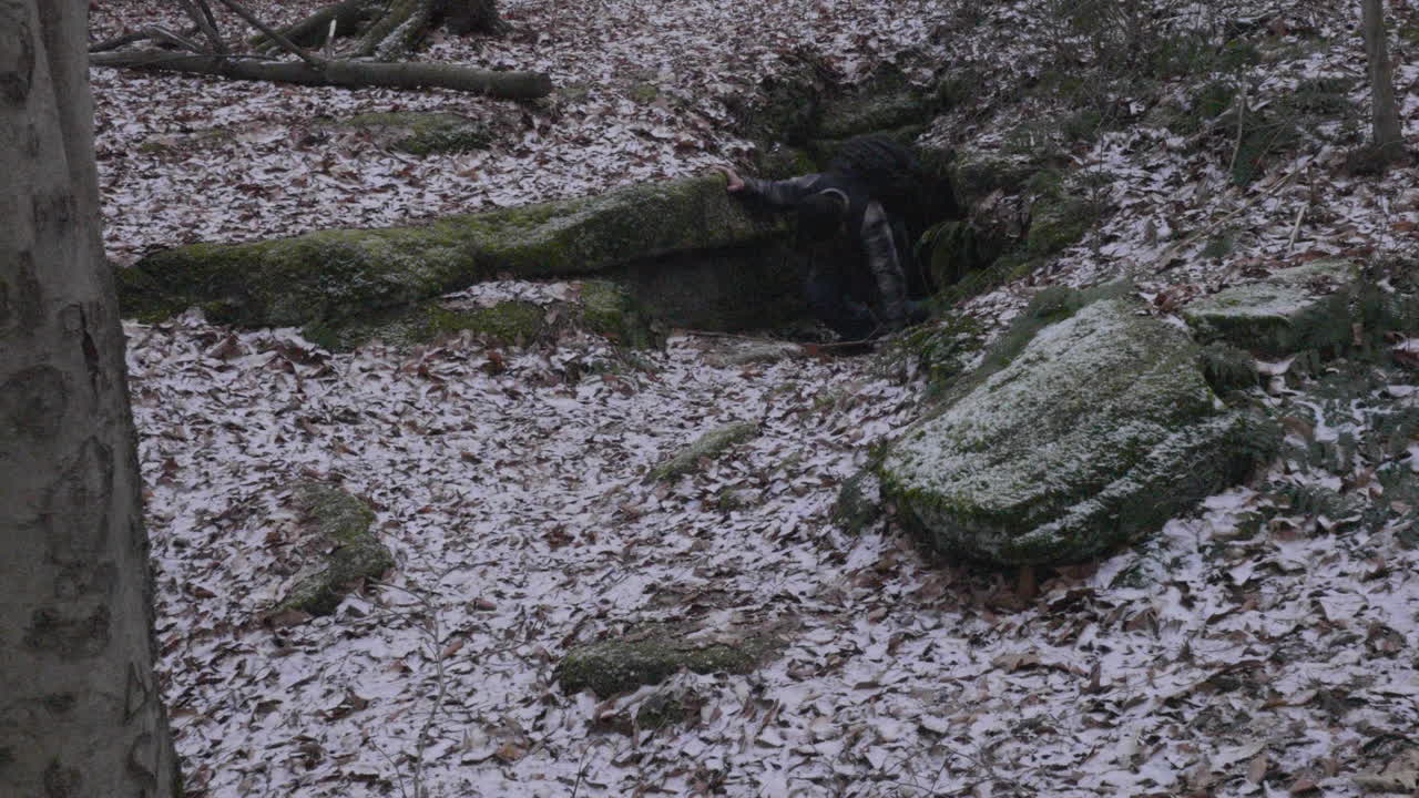 un excursionista sale de una cueva oscura escondida entre las rocas de mosey en el suelo del bosque cubierto de hojas en un día de invierno con un ligero polvo de nieve