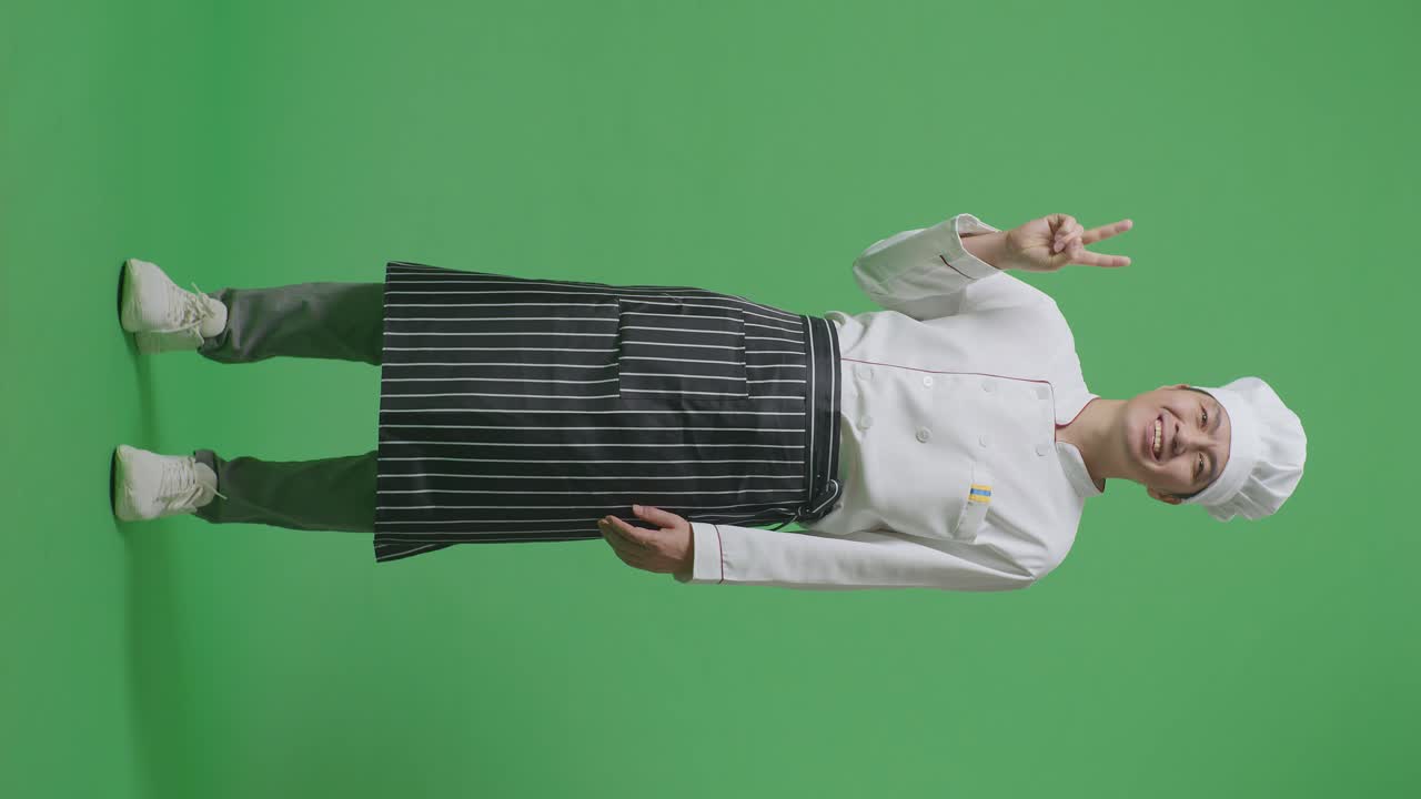 녹색 스크린 배경 스튜디오에 서있는 동안 카메라에 평화의 몸짓을 보여주고 미소 짓는 아시아 남자 셰프의 몸이 가득 차 있습니다.