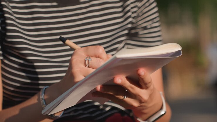 Nærbillede af hænder der skriver i spiralnotesbog udendørs, stribet skjorte, ringe, penstrøg fanger varmt sollys, fokuseret notatskrivning, tekstureret papir, afslappede smykker, eftermiddagsgadeatmosfære