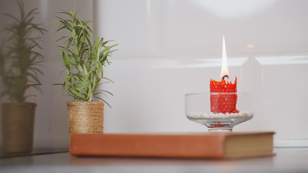 красная свеча горит в стеклянном держателе, заполненном белыми бусинками, помещенном рядом с декоративным зеленым растением в тканом горшке и закрытой книгой на столе напротив белой плиточной стены