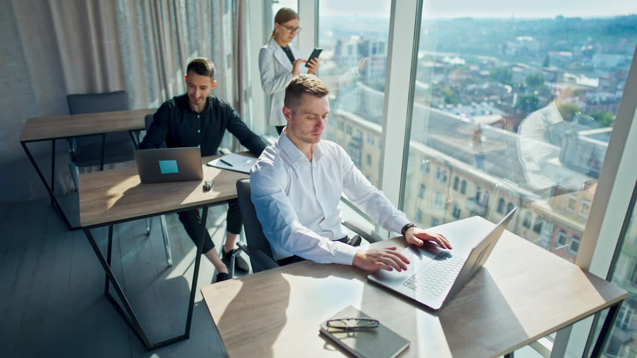 Male employees working at computers sitting at desks in sunny office. Female colleague stands near window using phone.