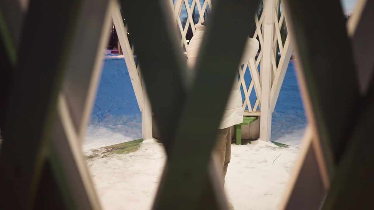 Through ornate lattice windows of winter gazebo dancer in quilted white coat and beanie wearing headphones moves fluidly with expressive gestures against snowy landscape lit by soft evening glow