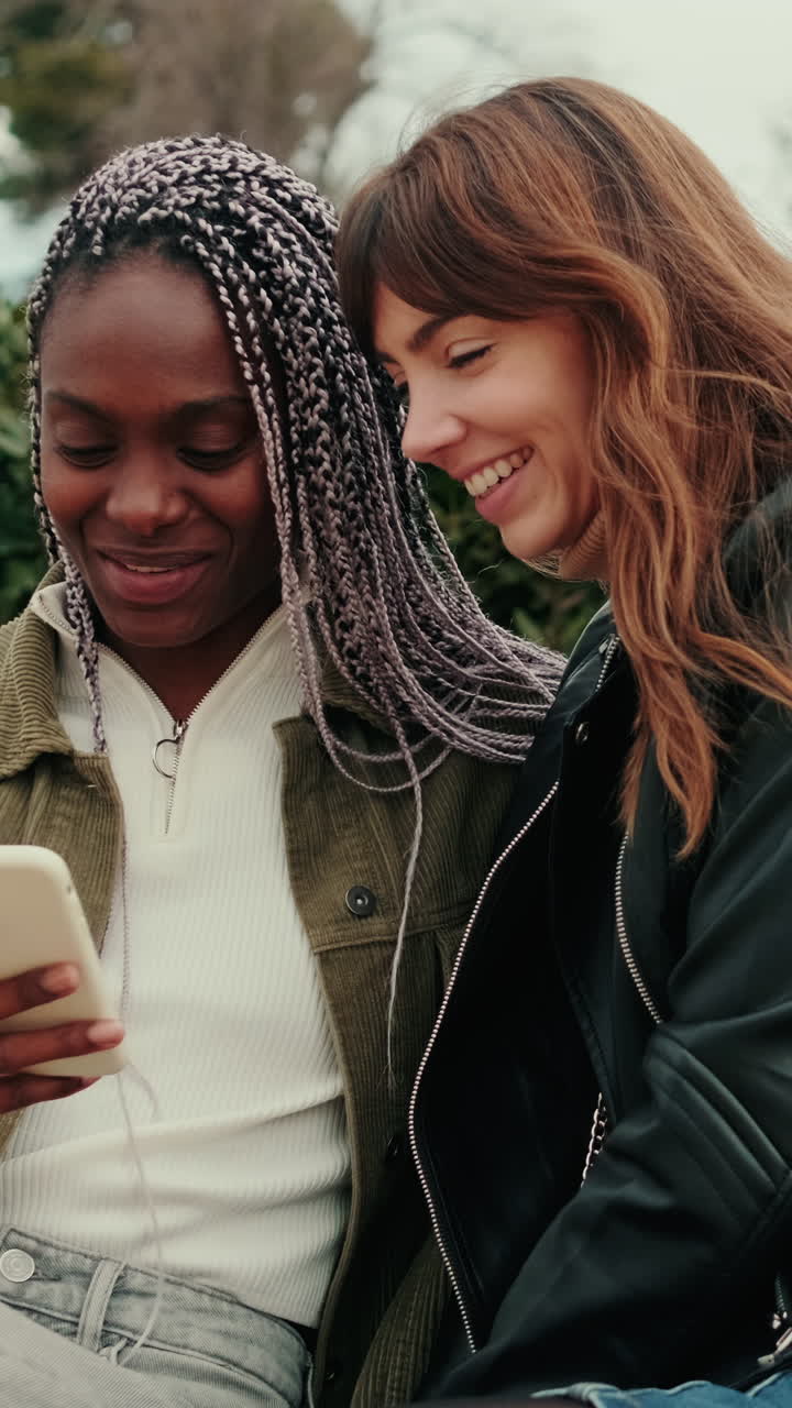 Two Young Women Sharing a Smartphone and Laughing Outdoors