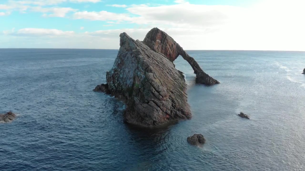 Drone stiger for at afsløre havbuen Bow Fiddle Rock, stille blåt hav, Skotland.
