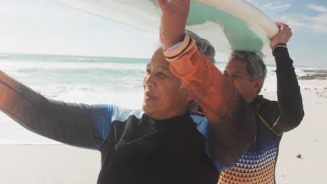 Happy-senior-hispanic-couple-walking-on-beach-with-surfboard-over-heads