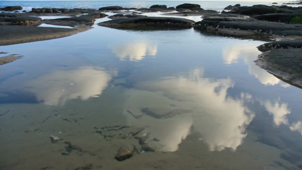 Reflecting shallow tidal pool showing shifting cumulus clouds on rocky shore, wind nudging surface