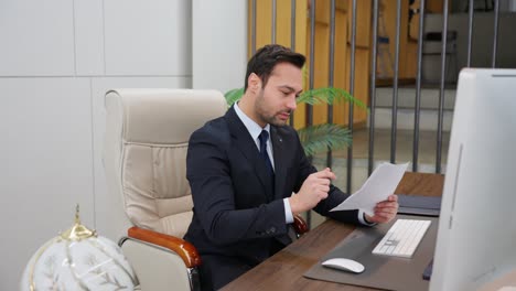busy thoughtful european businessman analyzing financial sheet documents checking data on computer while sitting at desk workplace in a business office. financier deals with financial report using pc