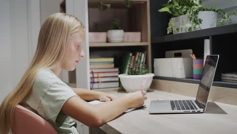 a teenage girl writes in a notebook, sits at a table near a laptop in her room. homeschooling concept