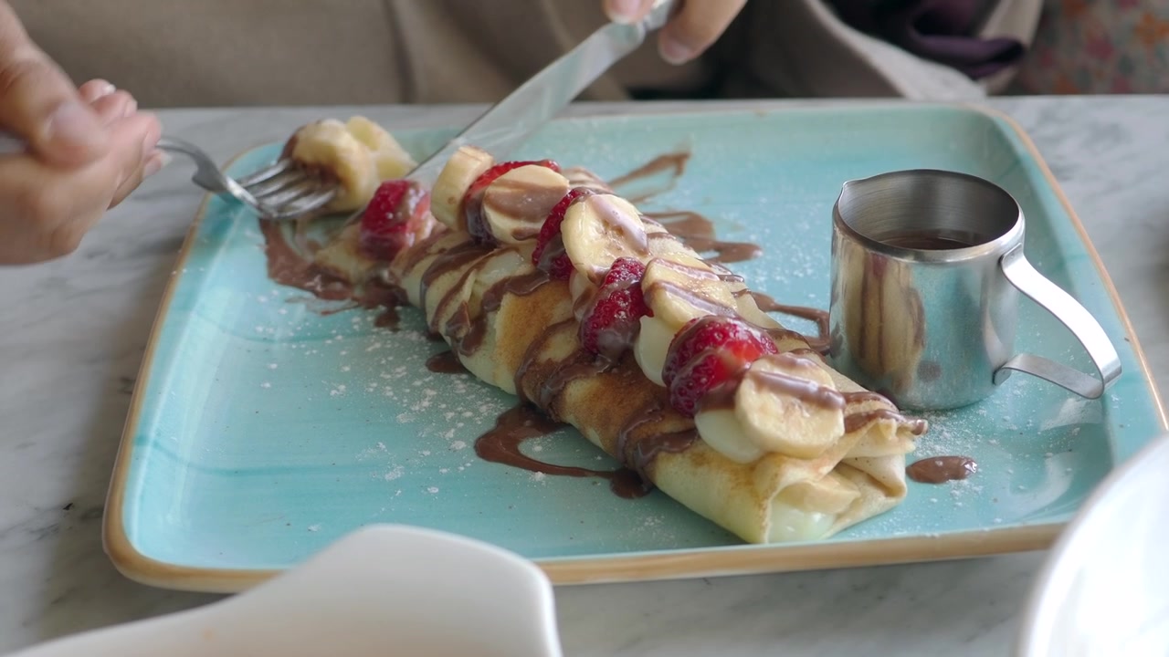 Persona che taglia e mangia una deliziosa crepe con banana, fragola e salsa al cioccolato