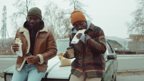 two african american men eating fast food and drinking coffee by car