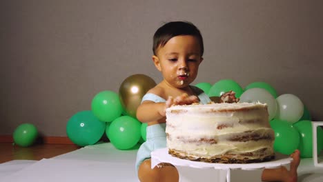 cute latin baby boy celebrating his 1st year birthday having fun biting a cake with green balloons in the background