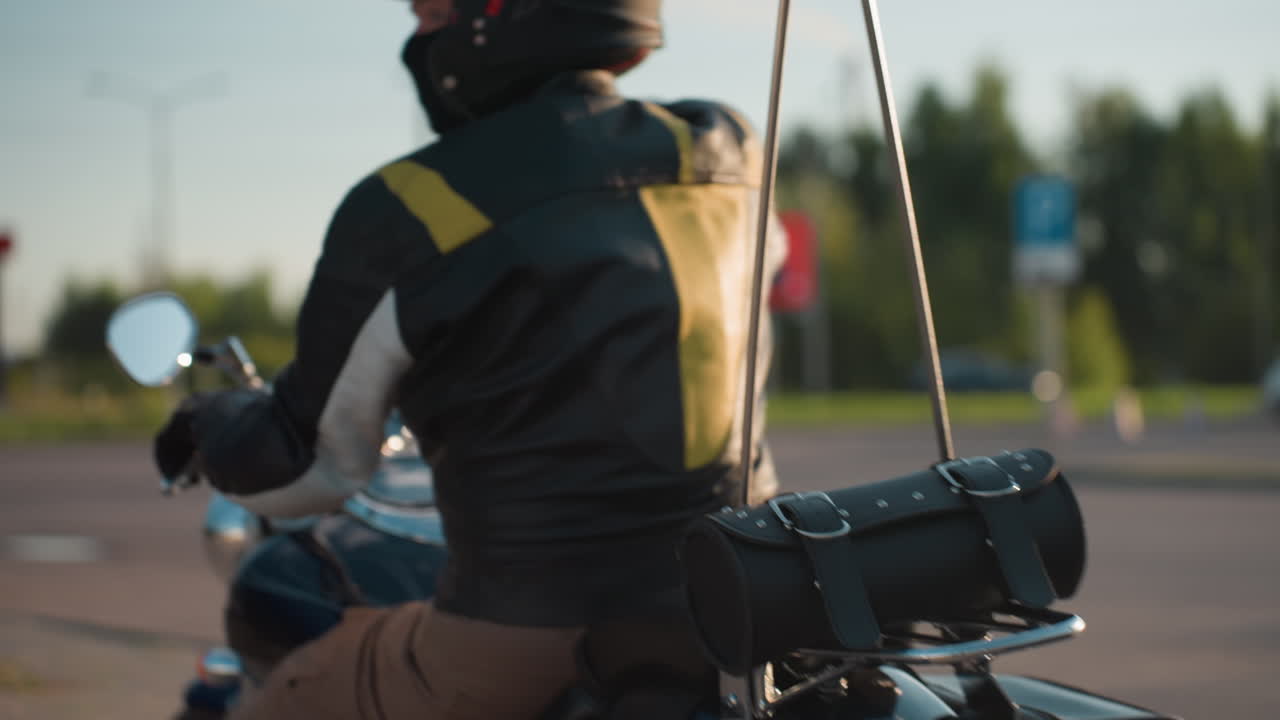Motorcycle rider wearing helmet and leather jacket drives off on road, side mirror reflecting light with blurred background of trees and street signs