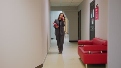 professional businesswoman walking through office corridor while talking on phone, holding folder. modern office environment, business attire, and stylish red sofa