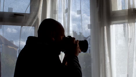 the individual peers through the window, camera in hand, framed by drawn curtains, evoking a sense of detective observation