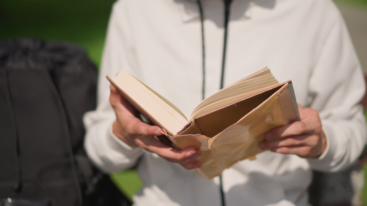 Un viajero caucásico lee un diario marrón al aire libre, sus manos hojean páginas texturizadas en un banco del parque iluminado por el sol, con una sudadera con capucha y una mochila a un lado, primer plano de la portada y el lomo desgastado, ambiente tranquilo y reflexivo