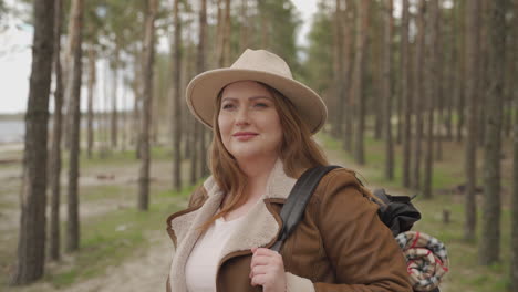 An-Adventurous-Red-Haired-Female-Camper-Wearing-A-Camel-Colored-Hat,-Backpack-And-Blanket-Looks-Around-Her-In-The-Forest