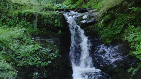 Aerial-drone-shot-zooms-out-to-reveal-a-small-waterfall-in-a-mountain-forest-adorned-with-lush-vegetation