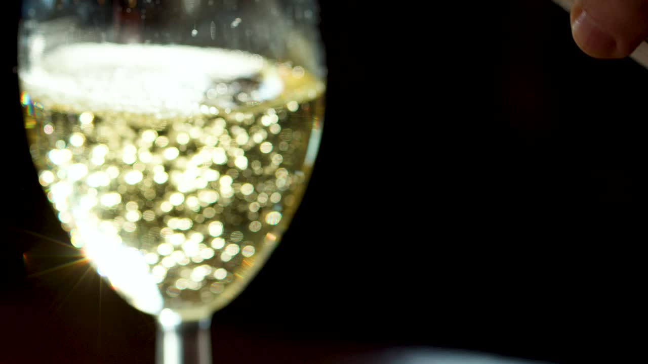 Sparkling wine poured into clear glass, bubbles rising, strong sunlight, dark background, static camera