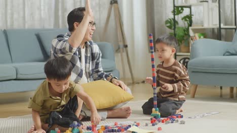 full body of asian father and sons assemble the construction set colorful plastic toy brick on a mat at home. the father clapping hands celebrating success with another son, giving high five gesture