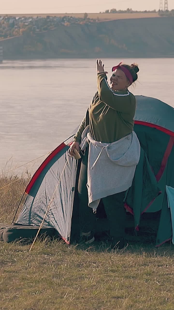 young plump woman gets out of blue tent drinks water from metal cup against pictorial calm river in morning slow motion
