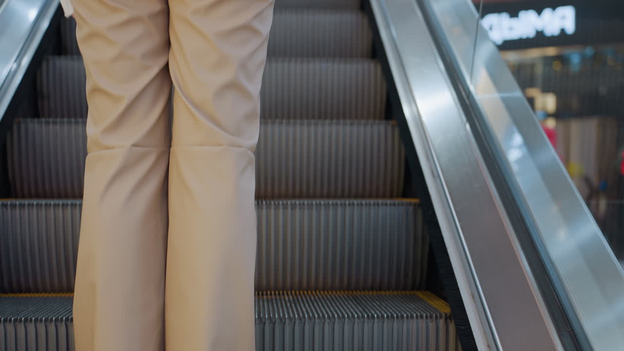 Rear view of woman with long brown hair in white sweater riding escalator in shopping mall, she gently holds escalator rail while moving upwards, with another person ahead